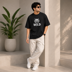 A man wearing a black graphic t-shirt with a white 'WILD' text and wolf design, paired with beige cargo pants and white sneakers, standing in a modern architectural setting with a potted plant.